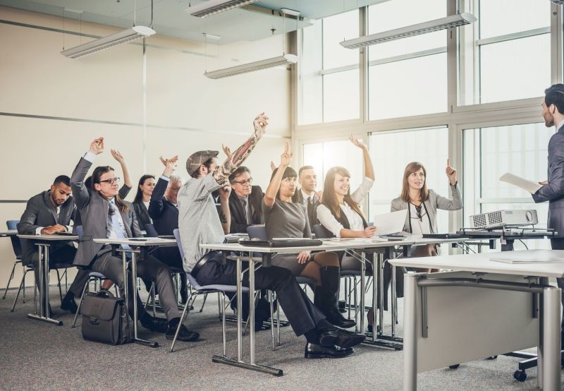 Multiple individuals in a training session sitting at desks with their hands raised and the leader at the front of the room.