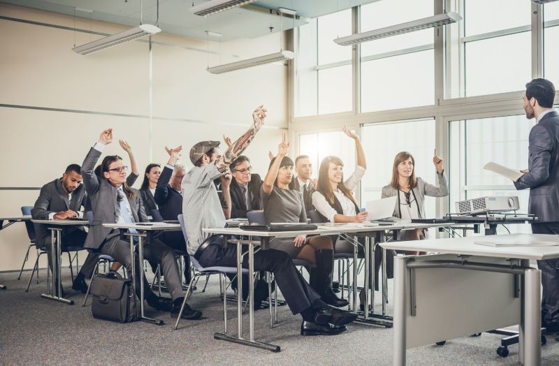 Multiple individuals in a training session sitting at desks with their hands raised and the leader at the front of the room.